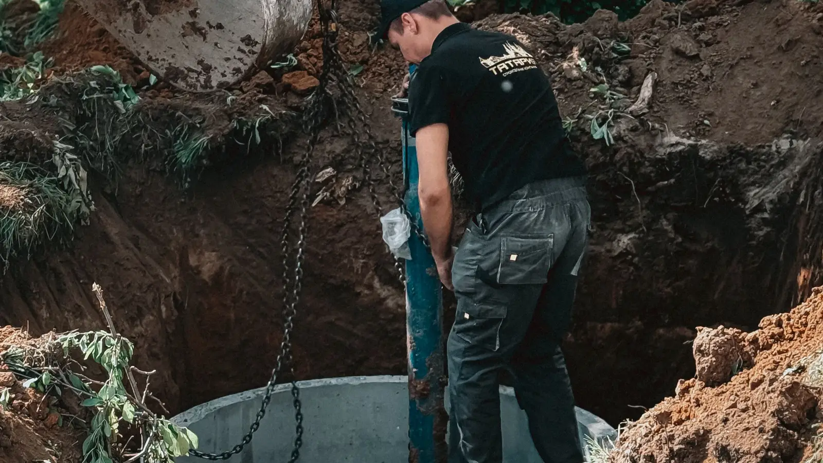 A worker fitting a concrete riser section into a freshly dug hole above a septic tank access port